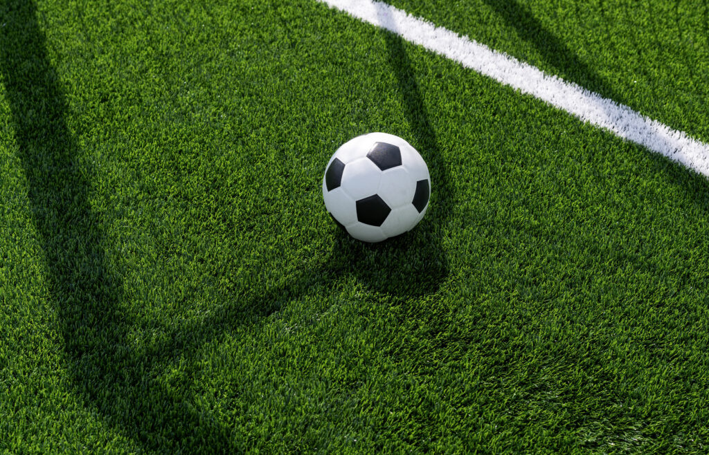 Black and white soccer ball on green turf near the goal line in the shadow of the goal.